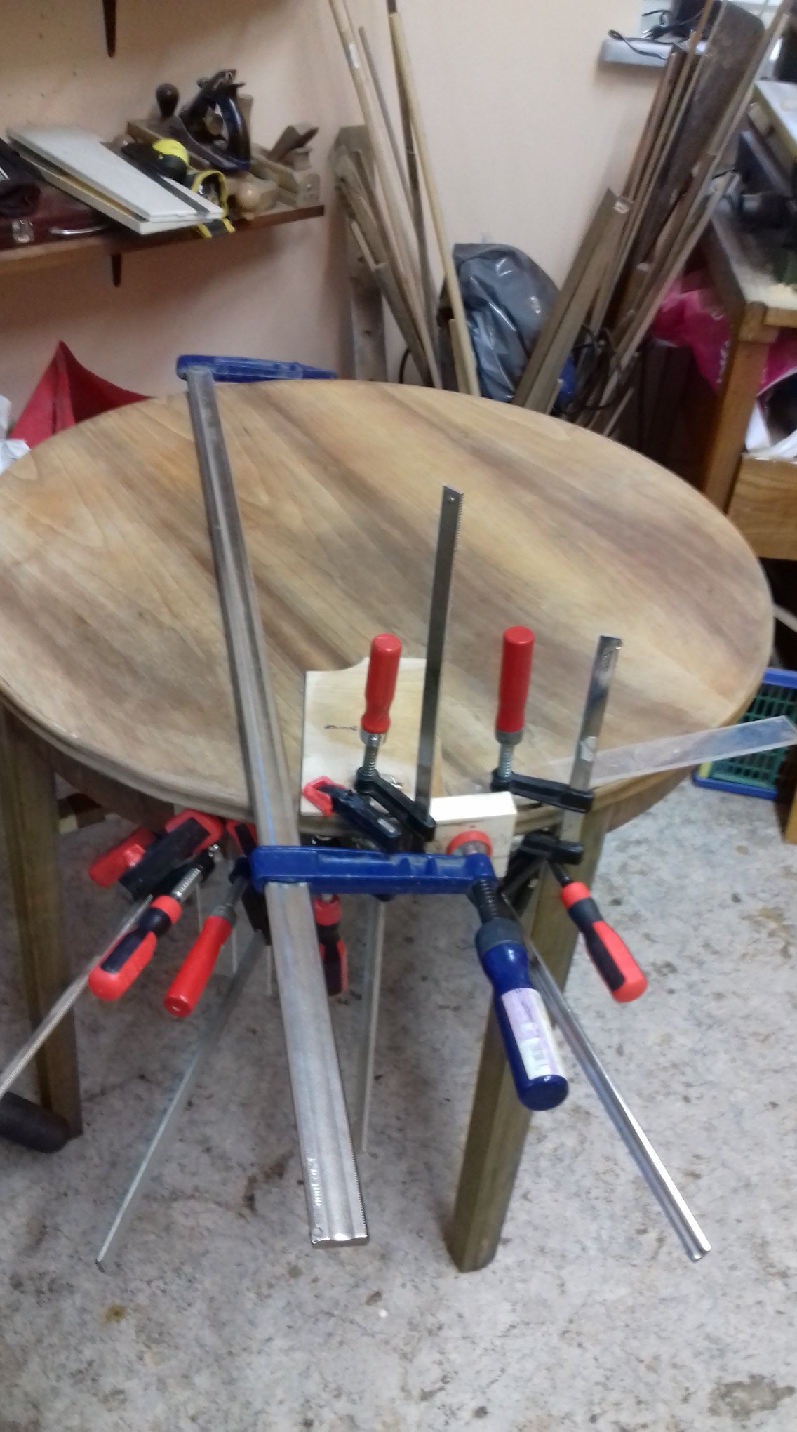 Okrągły, drewniany blat stołu w trakcie renowacji, zabezpieczony ściskami stolarskimi i listwami, w warsztacie.