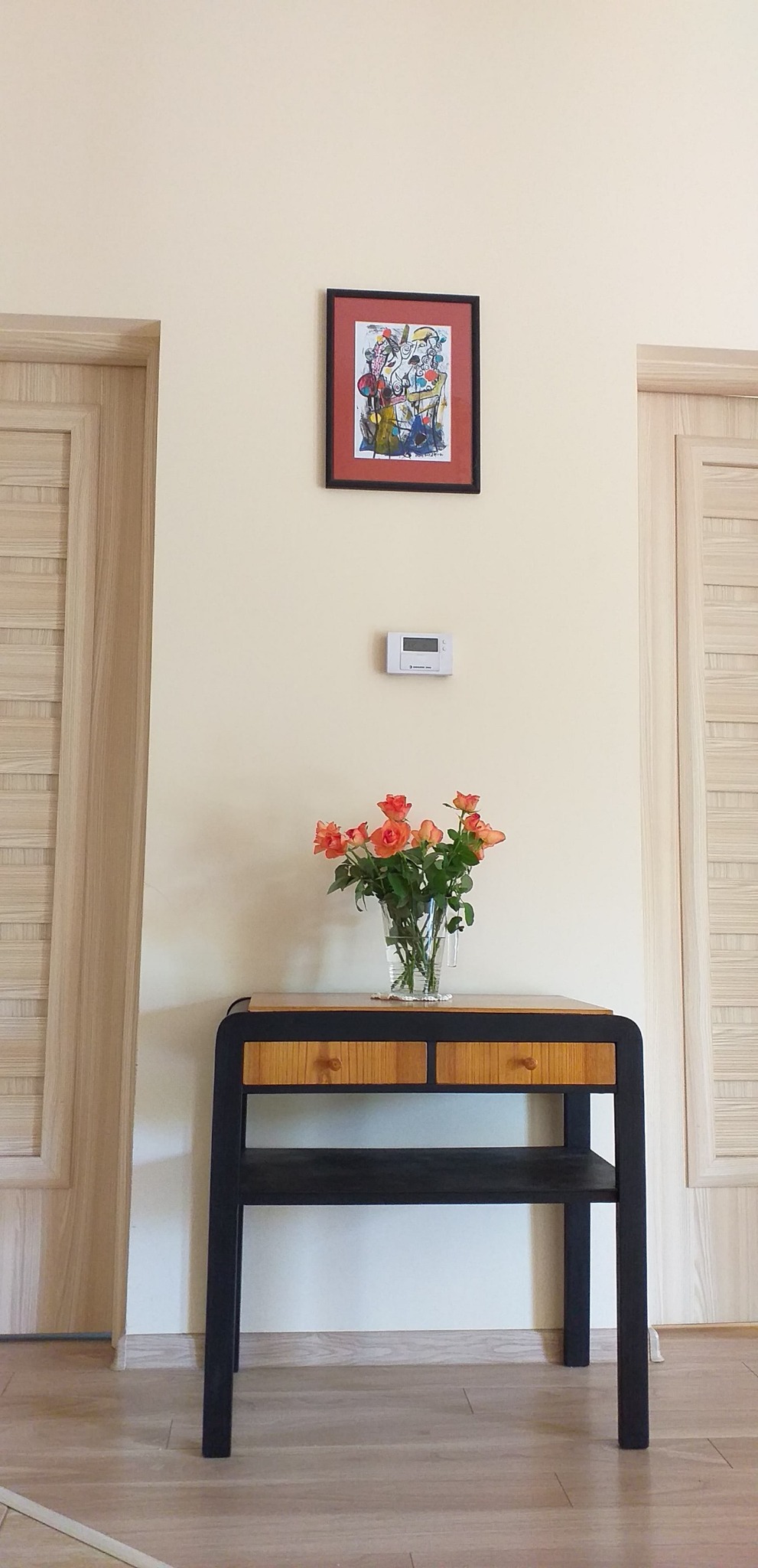 Wnętrze z odnowioną ścianą, ozdobioną abstrakcyjnym obrazem w czarnej ramie, termostatem i bukietem pomarańczowych róż w szklanym wazonie, stojącym na czarno-drewnianej konsoli z dwoma szufladami...