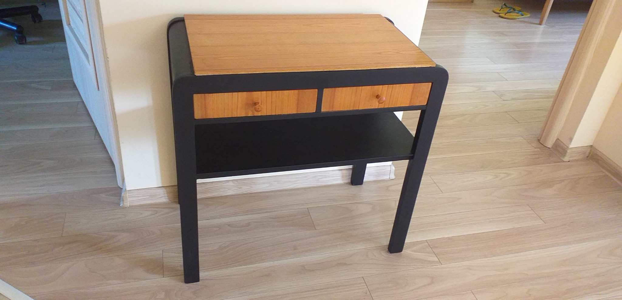 Odrestaurowany stolik nocny z dwoma drewnianymi szufladami i czarną ramą, ustawiony na drewnianej podłodze w jasnym wnętrzu.