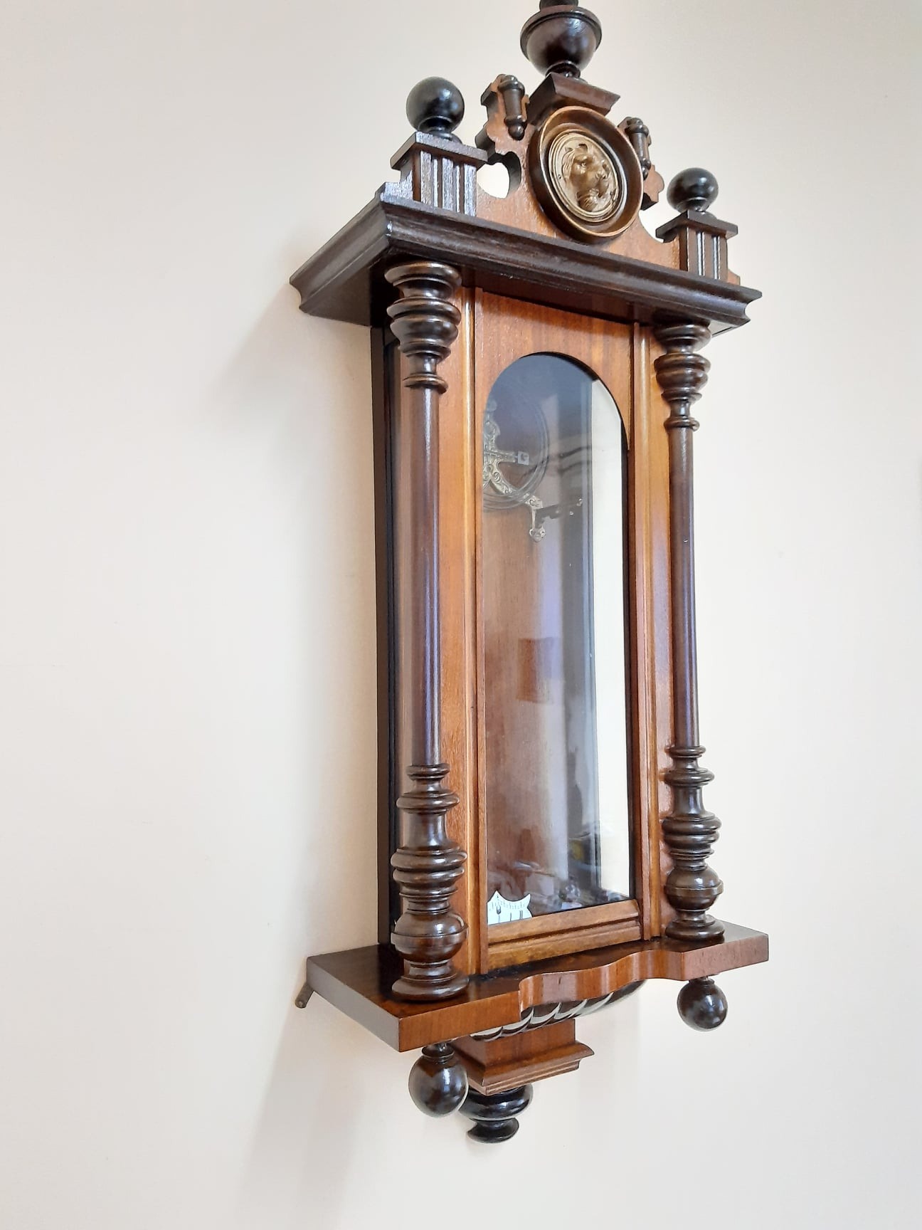 Wiszący, odrestaurowany zegar skrzynkowy w stylu secesyjnym z ciemnego drewna, z dekoracyjnym medalionem lwa na szczycie.