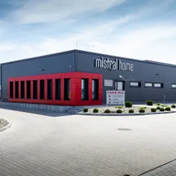 Nowoczesny budynek firmy Mistral Home z czerwonym akcentem architektonicznym, widoczny parking i zadbana zieleń, błękitne niebo w tle.
