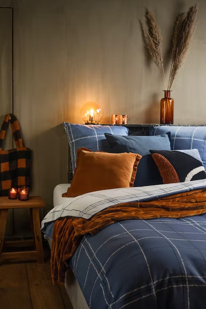 Nastrojowa sypialnia z pościelą w kratę, pomarańczowymi poduszkami, welurowym pledem i lampką nocną tworząca ciepły klimat.