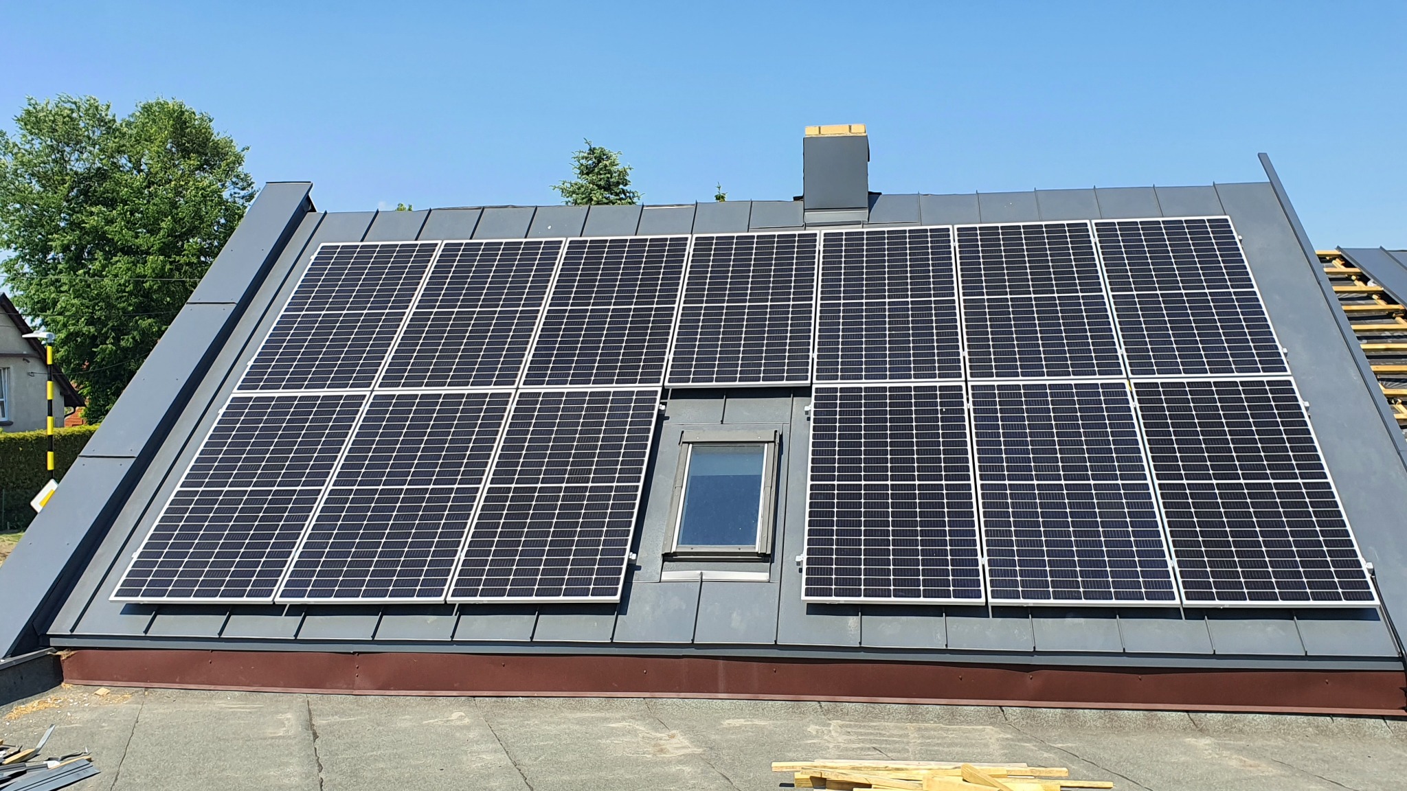 Panele słoneczne zamontowane na szarym dachu skośnym z oknem dachowym, widoczne fragmenty więźby dachowej i komin.