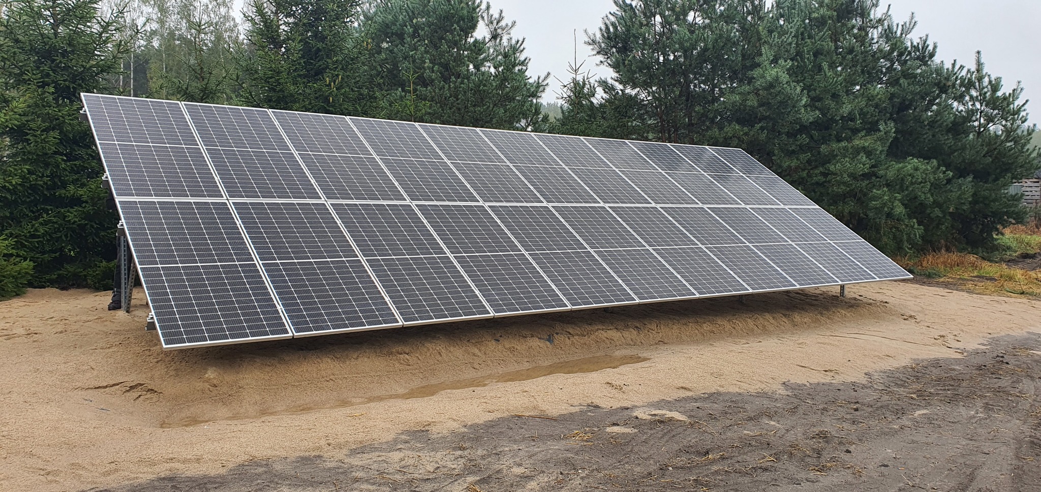 Instalacja paneli słonecznych na ziemi, widok na moduły fotowoltaiczne ustawione pod kątem na metalowej konstrukcji, w tle las iglasty w pochmurny dzień.
