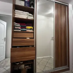 Wysoka, drewniana szafa wnękowa z przesuwnymi, lustrzanymi drzwiami, z otwartą częścią z półkami i szufladami, wypełniona ubraniami, ręcznikami i pudełkami, stojąca na podłodze wyłożonej płytkami.