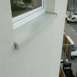 Szare wykończenie parapetu zewnętrznego, widok z góry na elewację budynku wielorodzinnego z balkonami i oknami.