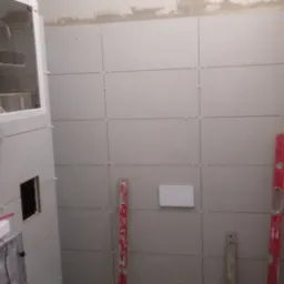 Łazienka w trakcie kładzenia płytek, widoczne poziomice, krzyżyki dystansowe i zabudowa stelaża podtynkowego z widocznym logo Synology.