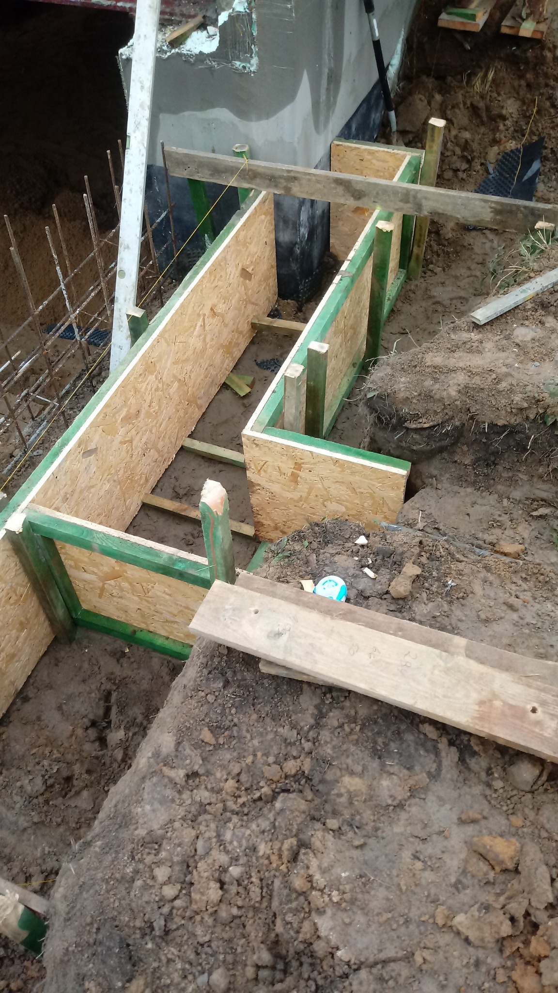 Szczegółowy widok wykopu z deskowaniem z płyty OSB, wzmocnionego zielonymi belkami, przygotowanego pod fundament; widoczne zbrojenie i izolacja pionowa na ścianie wykopu.