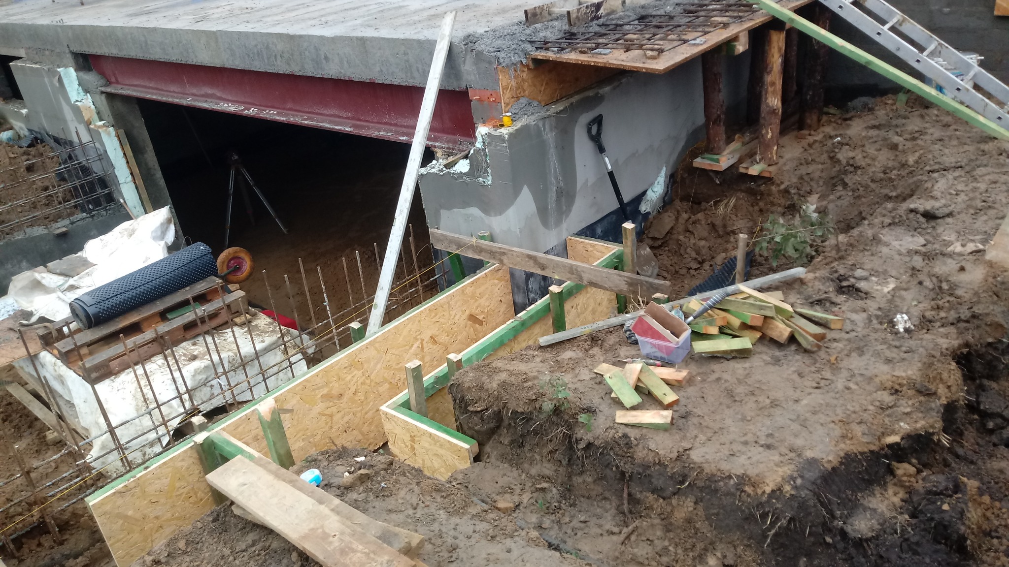 Budowa fundamentów domu: szalunek z płyty OSB, zbrojenie, częściowo wylany beton, widoczny przekrój ziemi, narzędzia budowlane i poziomica.