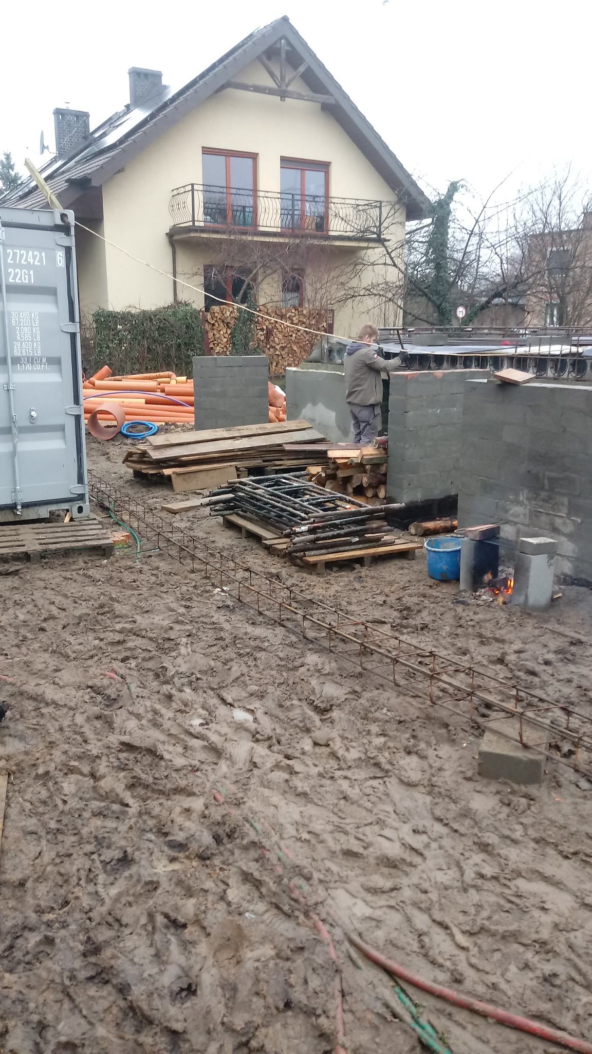 Budowa fundamentów domu: pracownik układa bloczki betonowe, widoczna stal zbrojeniowa, drewno, rury PCV oraz ogień w prowizorycznym palenisku na błotnistym terenie.
