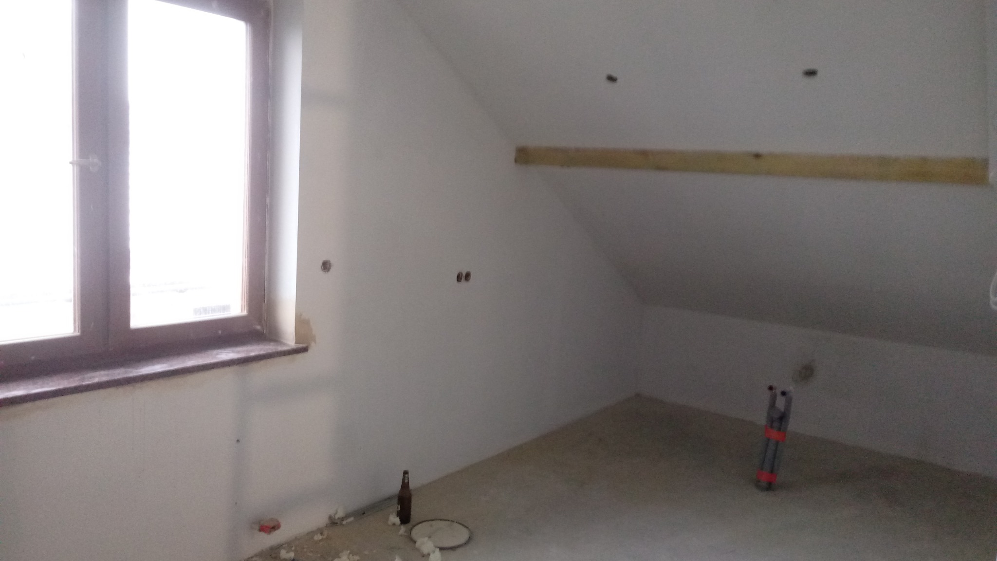 Poddasze w trakcie remontu z oknem, widoczne ślady prac, butelka po piwie i rozłożona drabina.