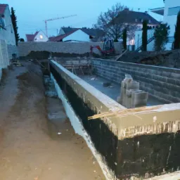 Fundamenty budynku w trakcie budowy, zabezpieczone czarną izolacją przeciwwilgociową; widoczny wykop, bloczki betonowe i mała koparka w tle.