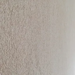 Ściana pokryta jasnobeżową, chropowatą strukturą gładzi, widoczna pod kątem, z delikatnym cieniowaniem w prawym górnym rogu.