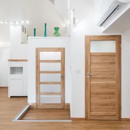 Jasne wnętrze poddasza z drewnianą podłogą, dwiema drewnianymi drzwiami z przeszkleniami, białymi ścianami, klimatyzacją i nowoczesnym oświetleniem.
