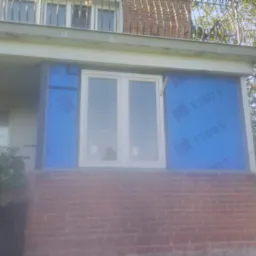 Montaż okna PCV na ceglanym balkonie, zabezpieczone niebieską folią ochronną PSB 1700 V, widoczne fragmenty roślinności i balustrady na wyższym poziomie budynku.