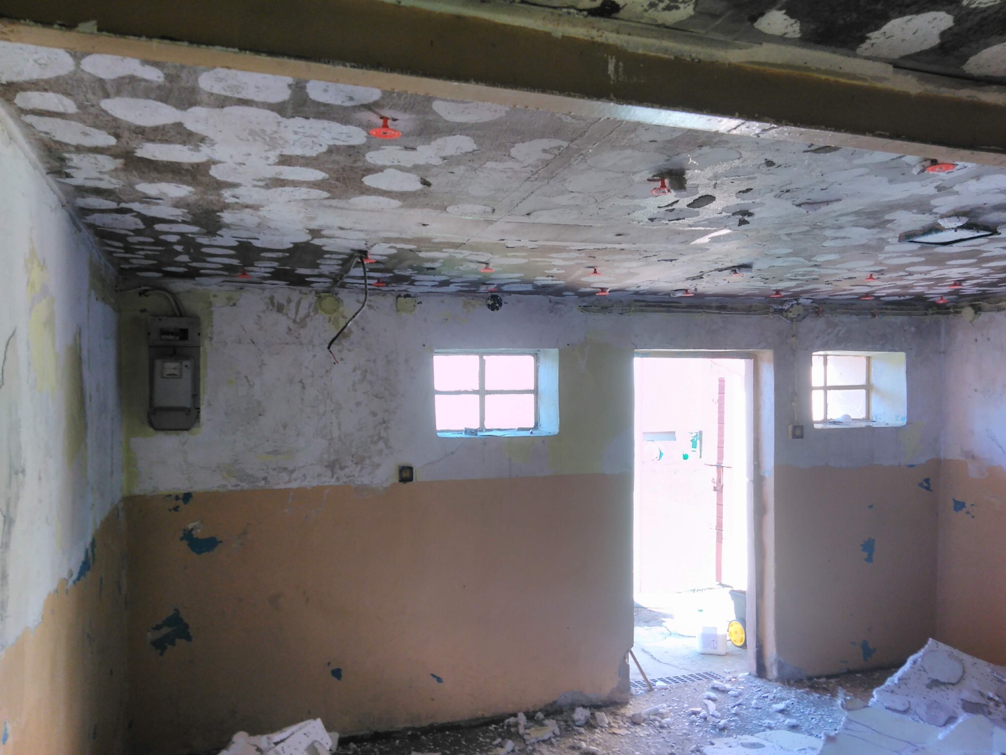 Pokój w trakcie przygotowań do remontu: surowy sufit z widocznymi punktami mocowania, ściany z resztkami farby i widocznym licznikiem elektrycznym, gruz na podłodze, okna z kratami.