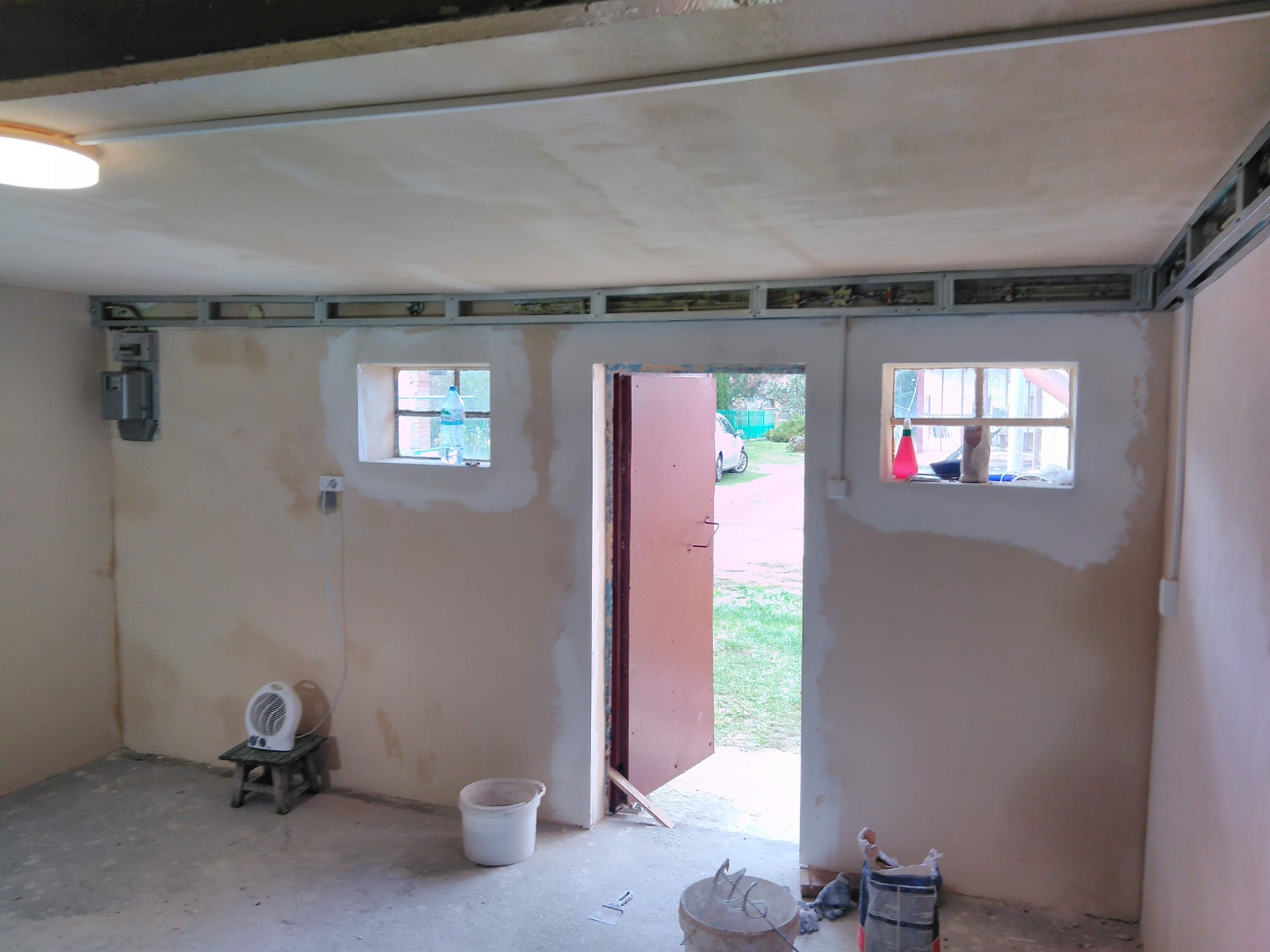 Wnętrze w trakcie prac wykończeniowych: ściany pokryte świeżo nałożoną gładzią, widoczne profile pod sufitem podwieszanym, narzędzia budowlane na podłodze, otwarte drzwi z widokiem na zewnątrz.