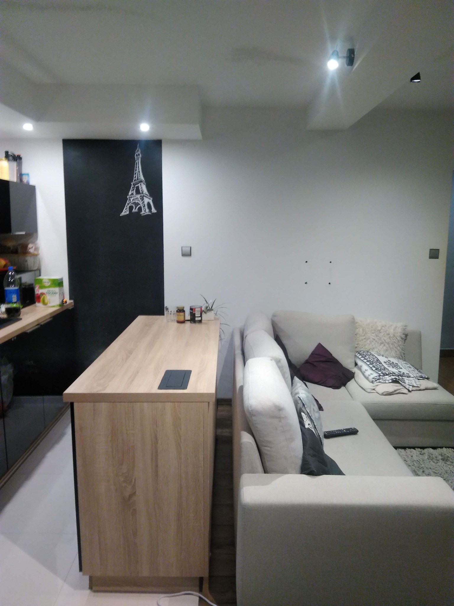 Jasne wnętrze salonu z aneksem kuchennym, drewniany blat wyspy kuchennej z ukrytymi gniazdkami, narożna sofa w odcieniach beżu, ściana z rysunkiem wieży Eiffla, nowoczesne oświetlenie punktowe.