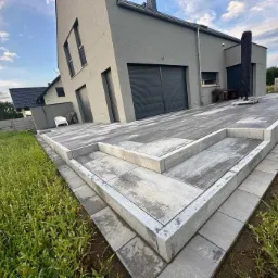 Nowoczesny taras z szarej kostki betonowej, z widocznymi schodami i minimalistycznym domem w tle, otoczony zielenią trawnika.