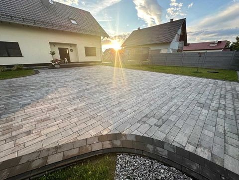 Ułożona szara kostka brukowa na podjeździe przed domem jednorodzinnym, widok z poziomu gruntu z dekoracyjnym obrzeżem z ciemnej kostki i kamieni, w tle zachodzące słońce.