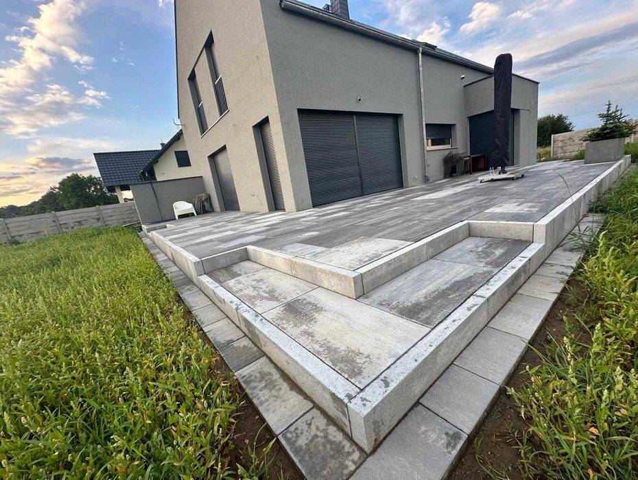 Nowoczesny taras z szarej kostki betonowej, z widocznymi schodami i minimalistycznym domem w tle, otoczony zielenią trawnika.