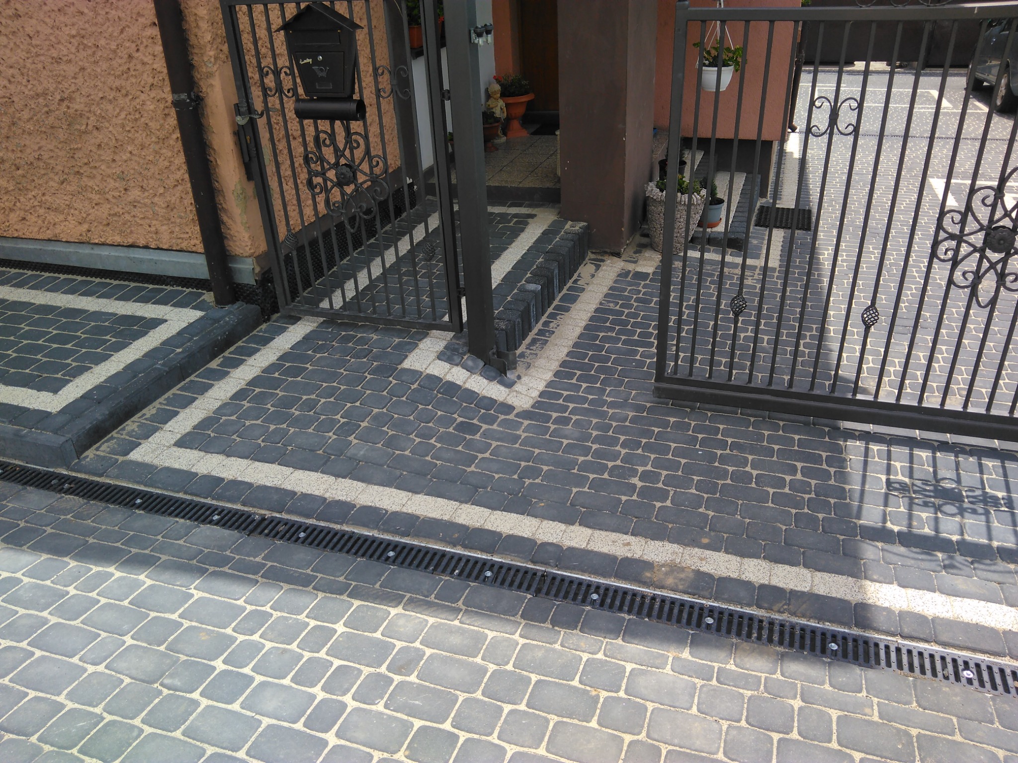 Ułożona kostka brukowa w odcieniach szarości i czerni z jasnymi akcentami przy posesji z kutą bramą i skrzynką na listy, widoczny system odprowadzania wody.