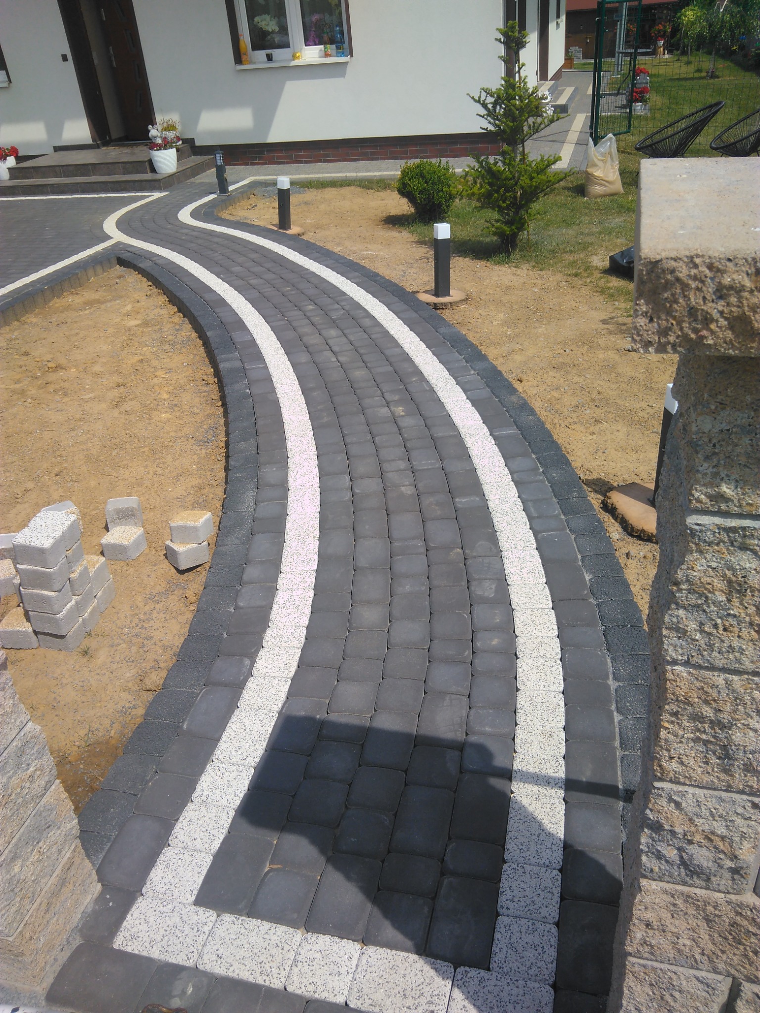 Ścieżka z ciemnoszarej kostki brukowej z białymi pasami, prowadząca do domu z jasną elewacją, widoczne ułożone kostki brukowe obok ścieżki.