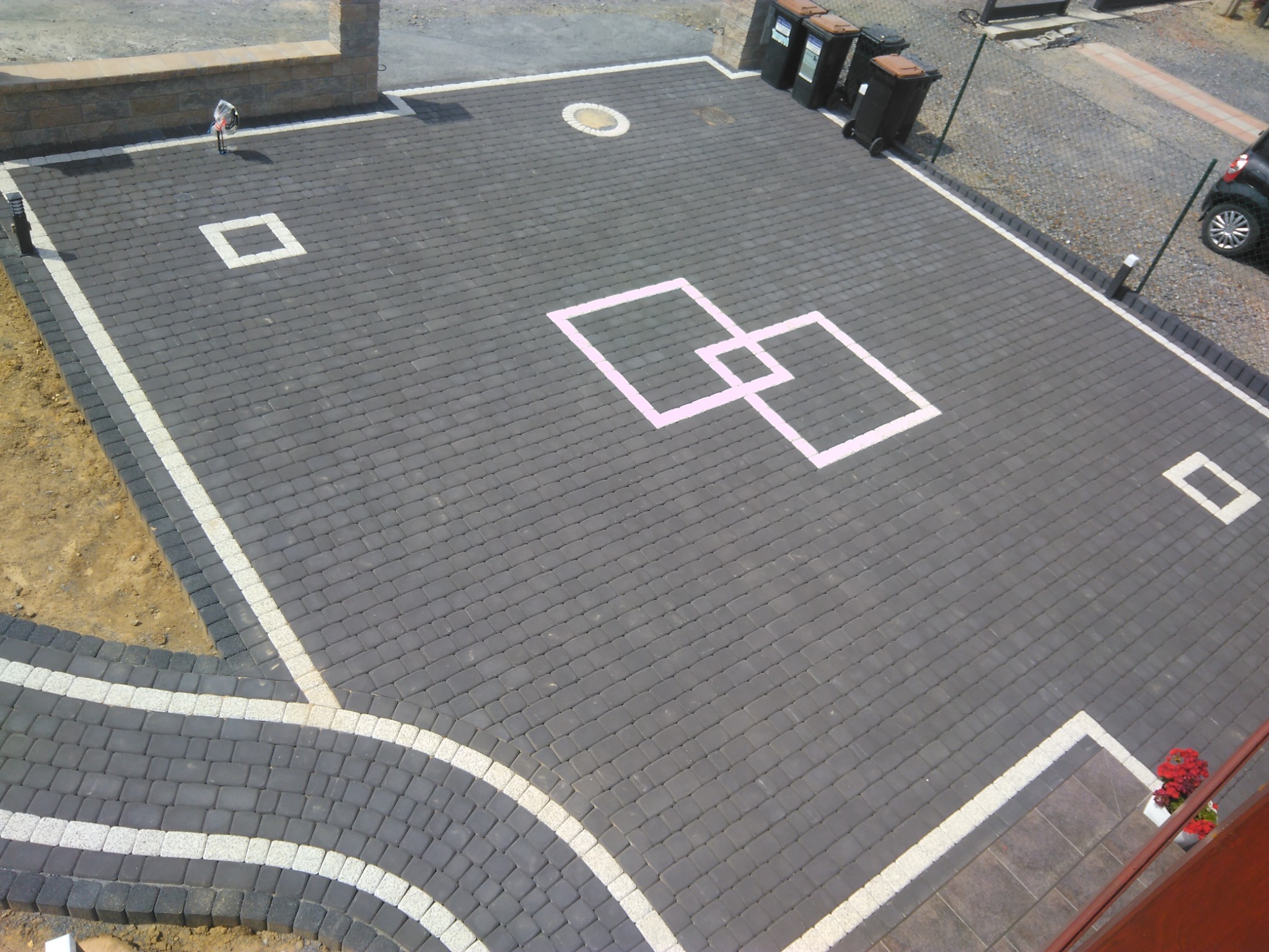 Rozległy podjazd z ciemnoszarej kostki brukowej, ozdobiony białymi obramowaniami i wzorem z różowych kwadratów, widok z góry.