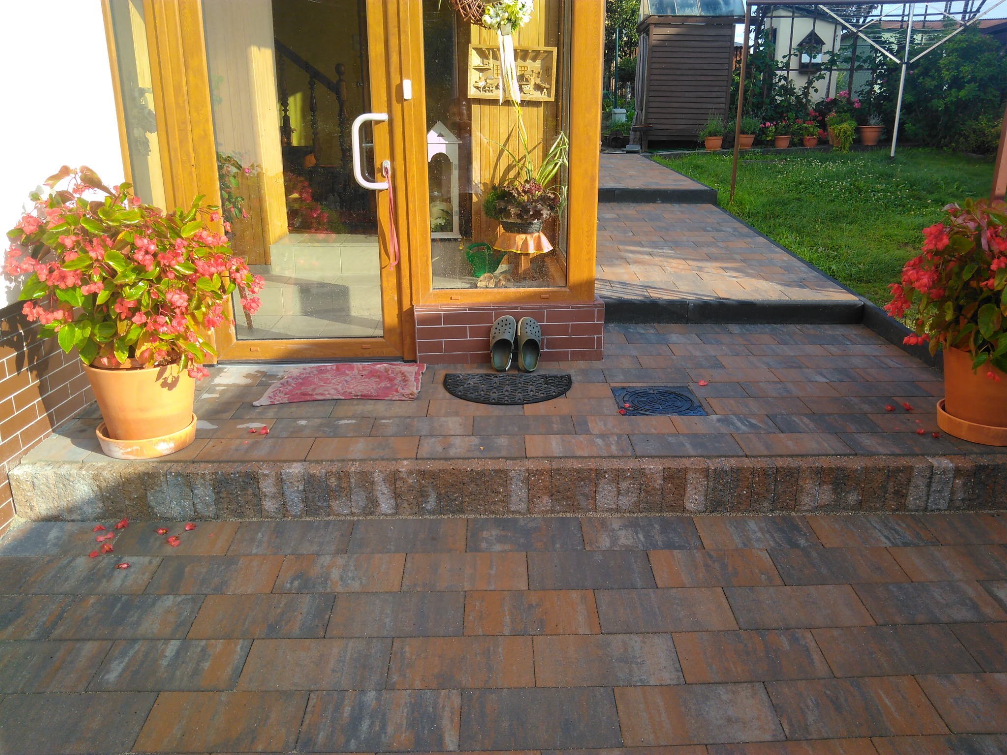 Brązowo-szara kostka brukowa prowadząca do drewnianych drzwi wejściowych, z donicami z czerwonymi kwiatami po bokach i parą klapek przed drzwiami.