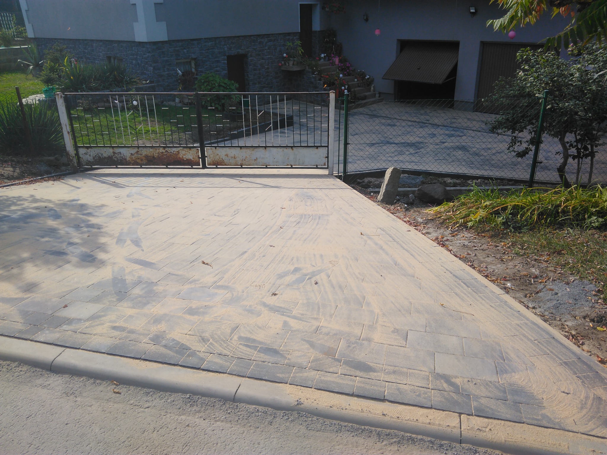 Nowa nawierzchnia z kostki brukowej prowadząca do posesji z otwartą bramą wjazdową, posypana piaskiem, widoczne ślady stóp.