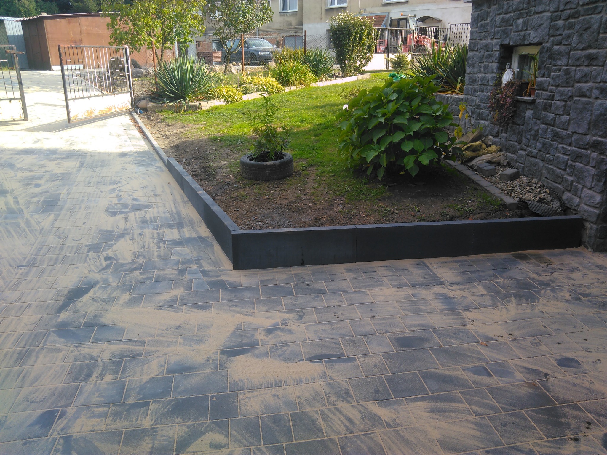 Szare kostki brukowe ułożone na podjeździe, z widocznymi śladami piasku, obok rabata z roślinnością i budynek z kamienną elewacją.