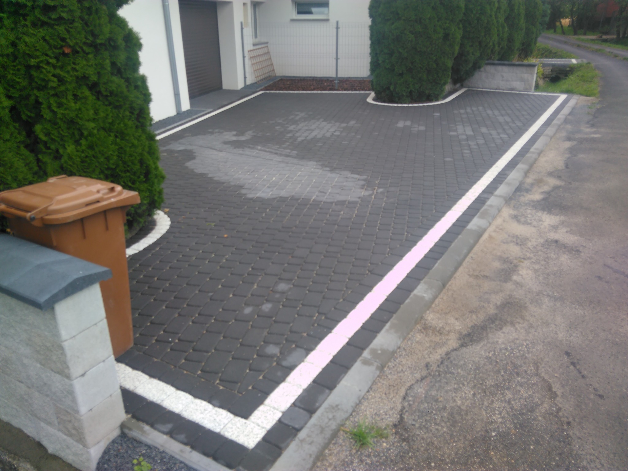 Ułożona ciemnoszara kostka brukowa na podjeździe z białym obramowaniem, widoczny brązowy pojemnik na śmieci i fragment ogrodu z tujami.