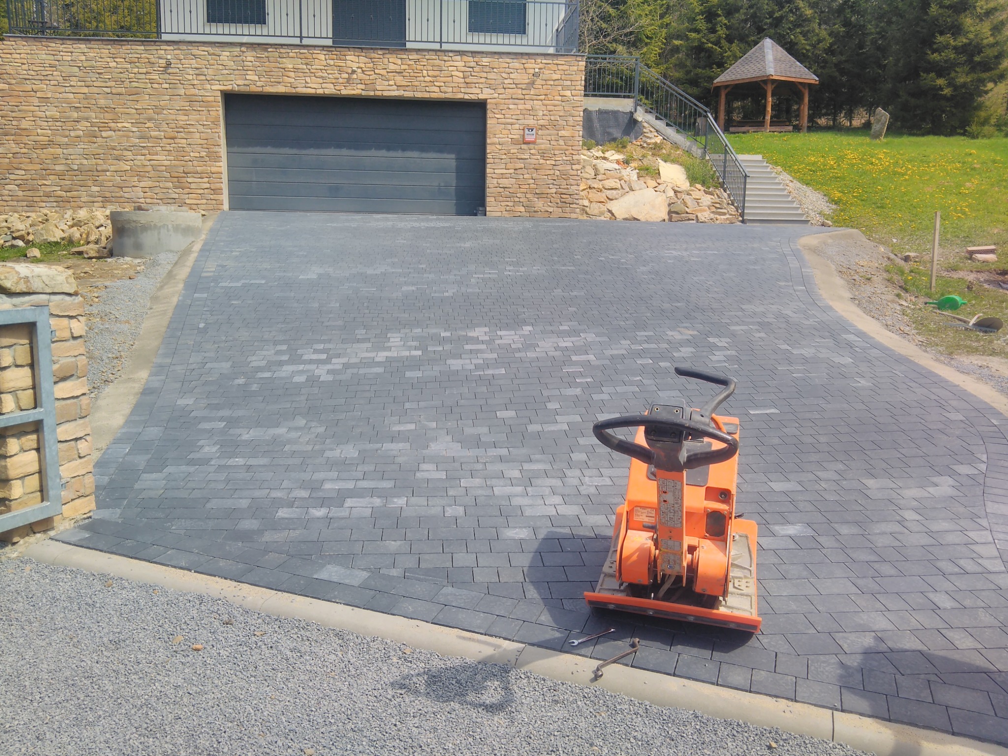Nowa, szara nawierzchnia z kostki brukowej przed domem z garażem; pomarańczowa zagęszczarka wibracyjna stoi na kostce, obok leżą klucze; w tle widoczna altana i schody.