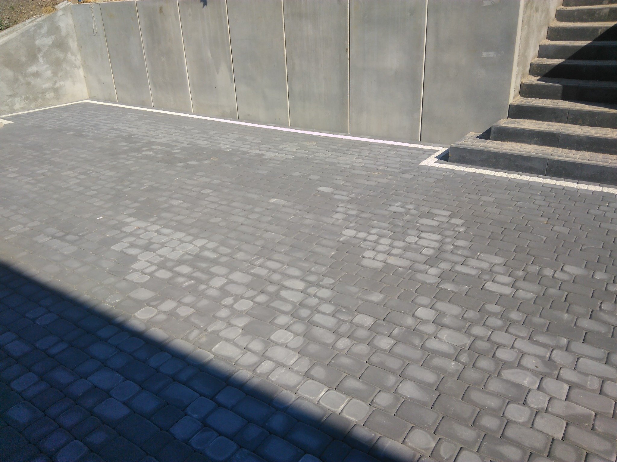 Szara kostka brukowa ułożona na podjeździe, przylegająca do betonowego muru oporowego i schodów, częściowo zacieniona.