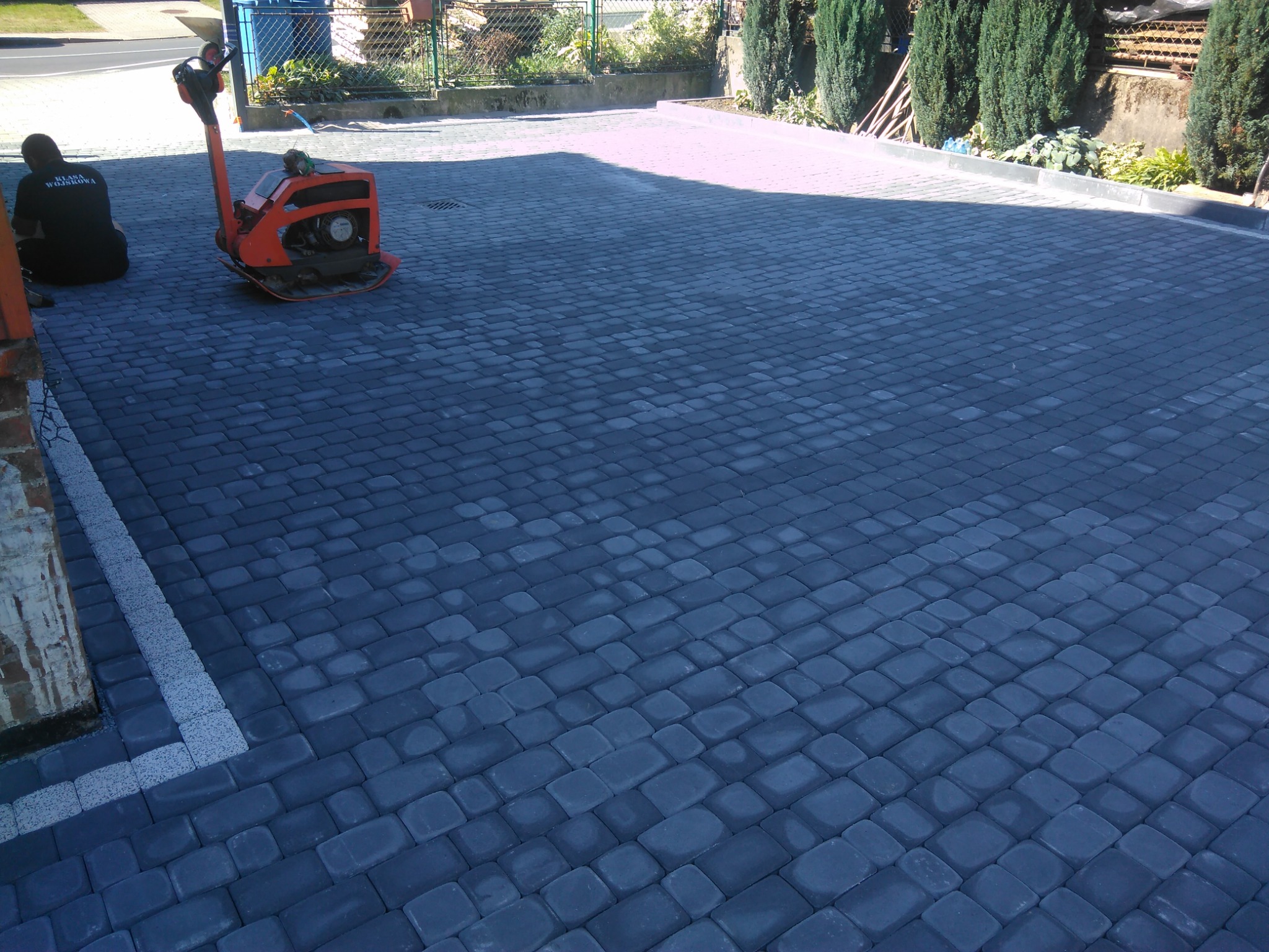 Świeżo ułożona, ciemnoszara kostka brukowa na posesji, z widoczną zagęszczarką w tle i fragmentem muru z jasną obwódką. Pracownik w ciemnej odzieży siedzi obok. Tło stanowi zieleń ogrodu i ogrodzenie.