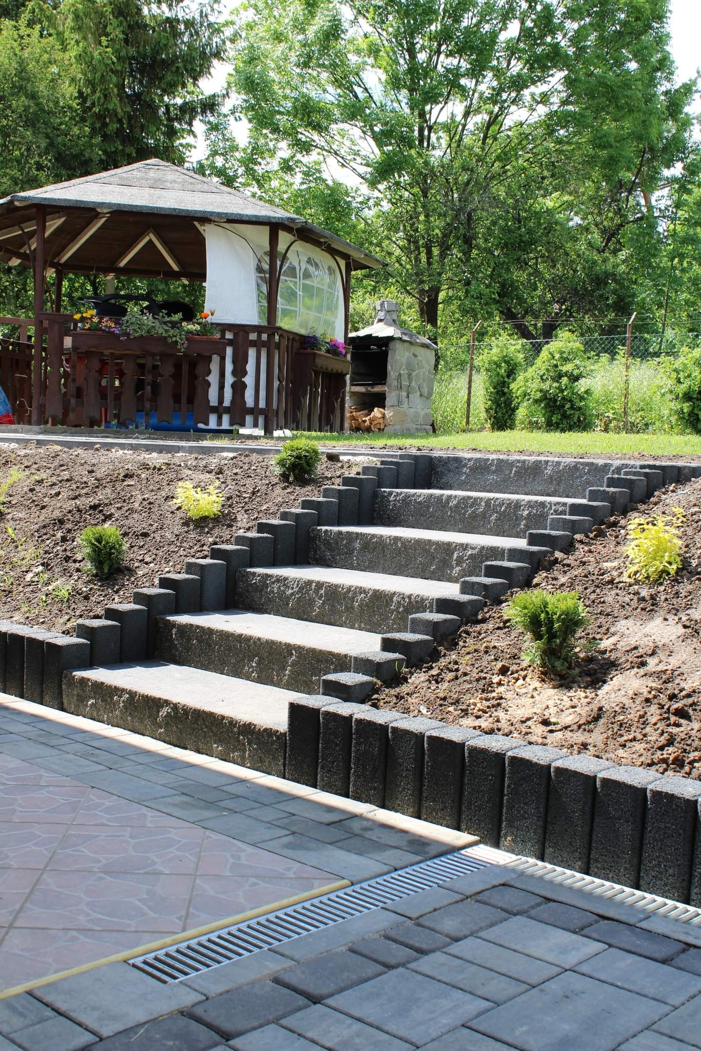 Schody ogrodowe z kostki betonowej prowadzące do altany, obrzeża z ciemnej kostki, widoczna kratka odpływowa i fragment brukowanej nawierzchni.