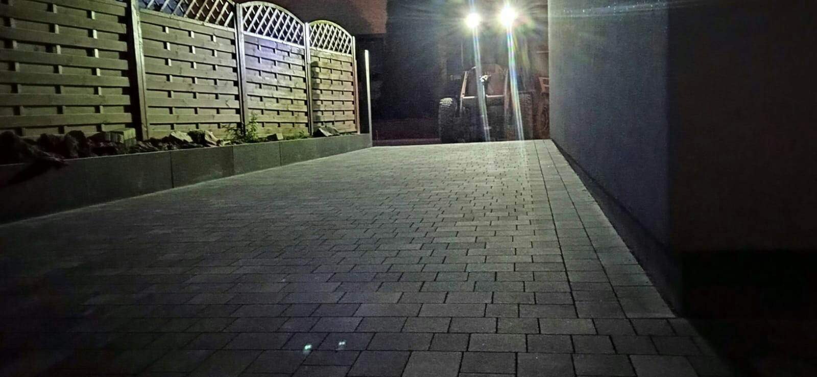 Ułożona kostka brukowa wzdłuż ogrodzenia, oświetlona przez reflektory maszyny budowlanej w tle.