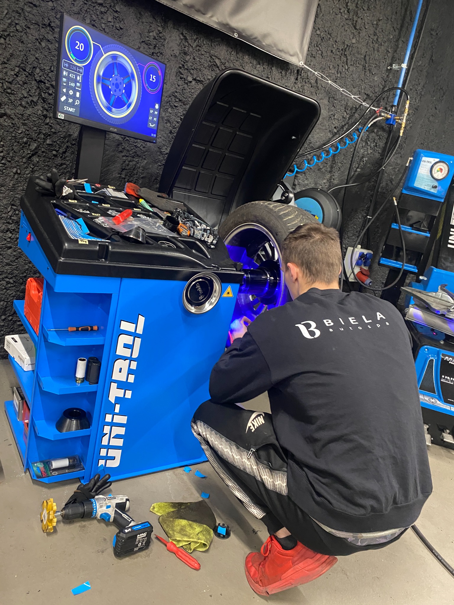 Mężczyzna w czarnej bluzie z logo 'BIELA autospa' kuca przy niebieskiej maszynie Uni-Trol do wyważania kół, z podświetlonym na niebiesko kołem, na podłodze leżą narzędzia, a na monitorze widoczne...