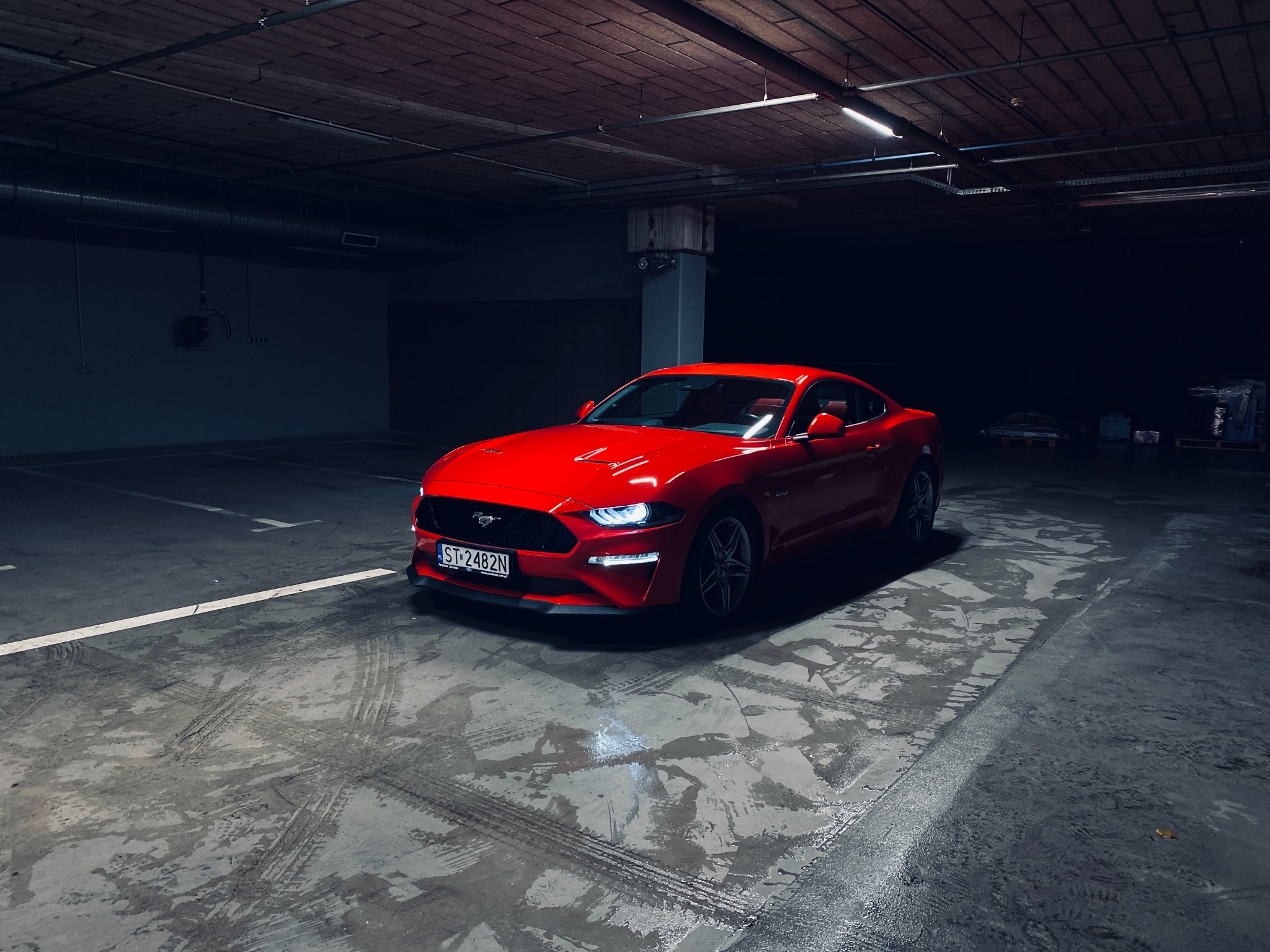 Czerwony Ford Mustang zaparkowany na betonowym parkingu podziemnym, widoczne ślady opon i mokra nawierzchnia.