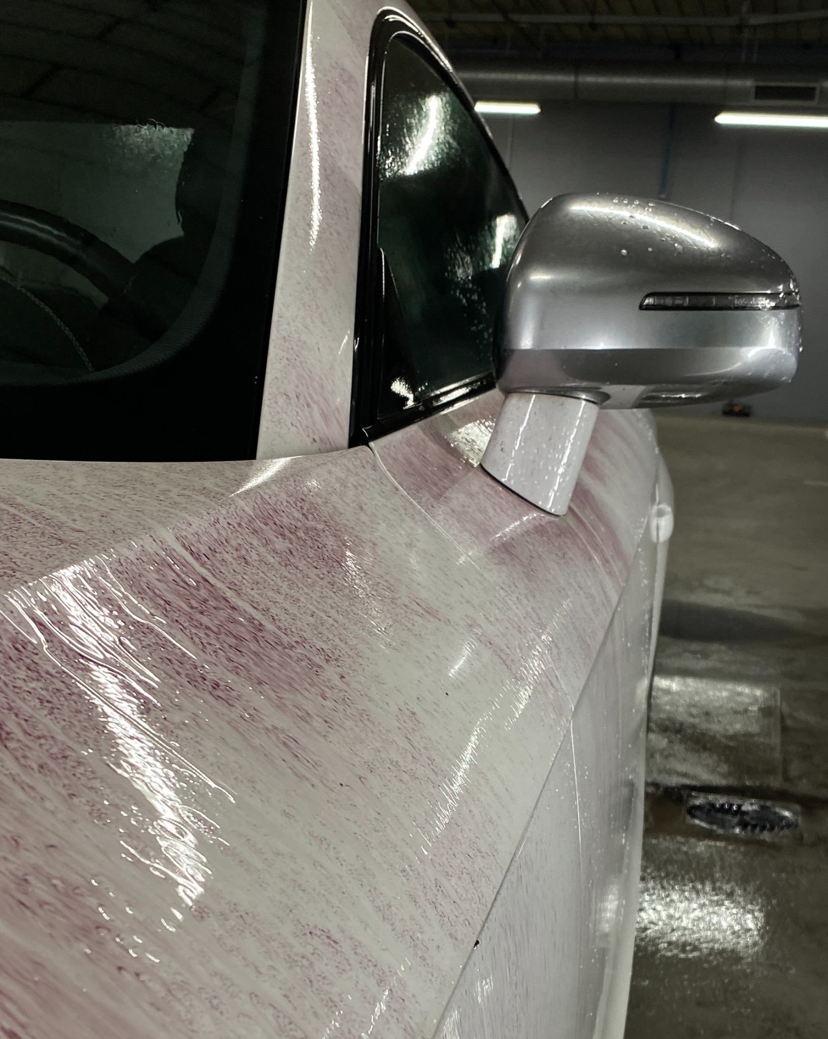 Biały samochód pokryty aktywną pianą o różowym zabarwieniu podczas mycia w myjni, widok z boku na lusterko i przednią szybę.