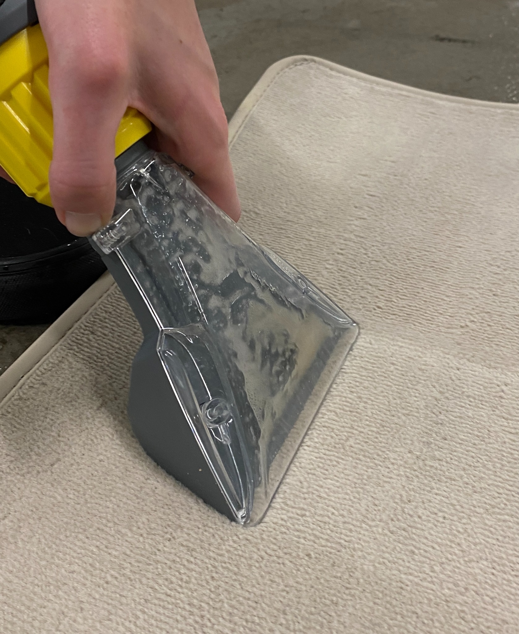 Czyszczenie beżowej tkaniny dywanowej za pomocą ekstraktora z przezroczystą dyszą i żółtą rączką, widoczne drobne zabrudzenia w dyszy.