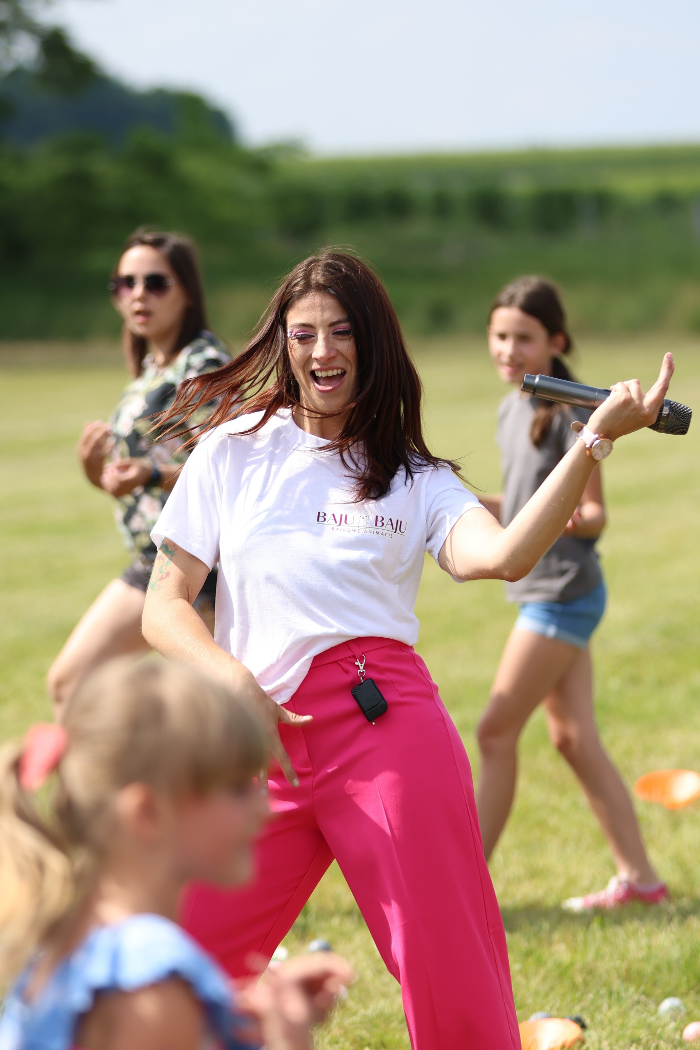 Animator w różowych spodniach i białej koszulce z logo firmy 'BAJU BAJU' prowadzi animacje dla dzieci na trawie, trzymając mikrofon w uniesionej ręce.