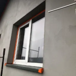 Ocieplanie elewacji budynku z widocznymi oknami zabezpieczonymi pomarańczową taśmą malarską, nałożona siatka zbrojąca przed tynkowaniem.