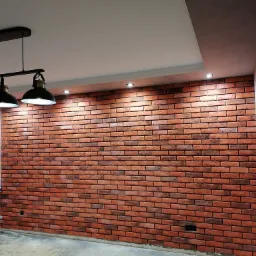 Ściana wykończona czerwoną cegłą klinkierową, oświetlona trzema lampami wiszącymi w stylu industrialnym i punktowymi reflektorami w suficie podwieszanym.
