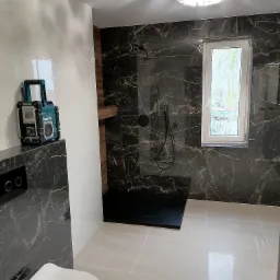 Nowoczesna łazienka z czarnymi marmurkowymi płytkami na ścianach, białymi płytkami na podłodze i ścianie, kabiną prysznicową typu walk-in z czarną armaturą, oraz radiem budowlanym Makita stojącym...
