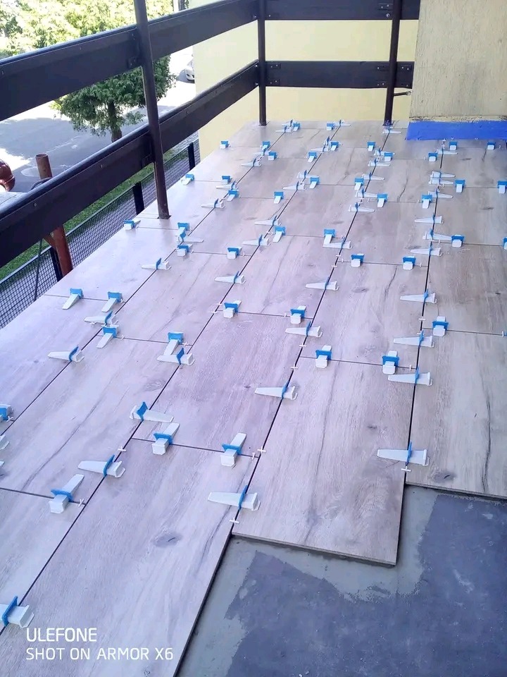 Montaż płytek imitujących drewno na balkonie z użyciem systemu poziomowania, widoczne krzyżyki i kliny, częściowo ukończona powierzchnia.