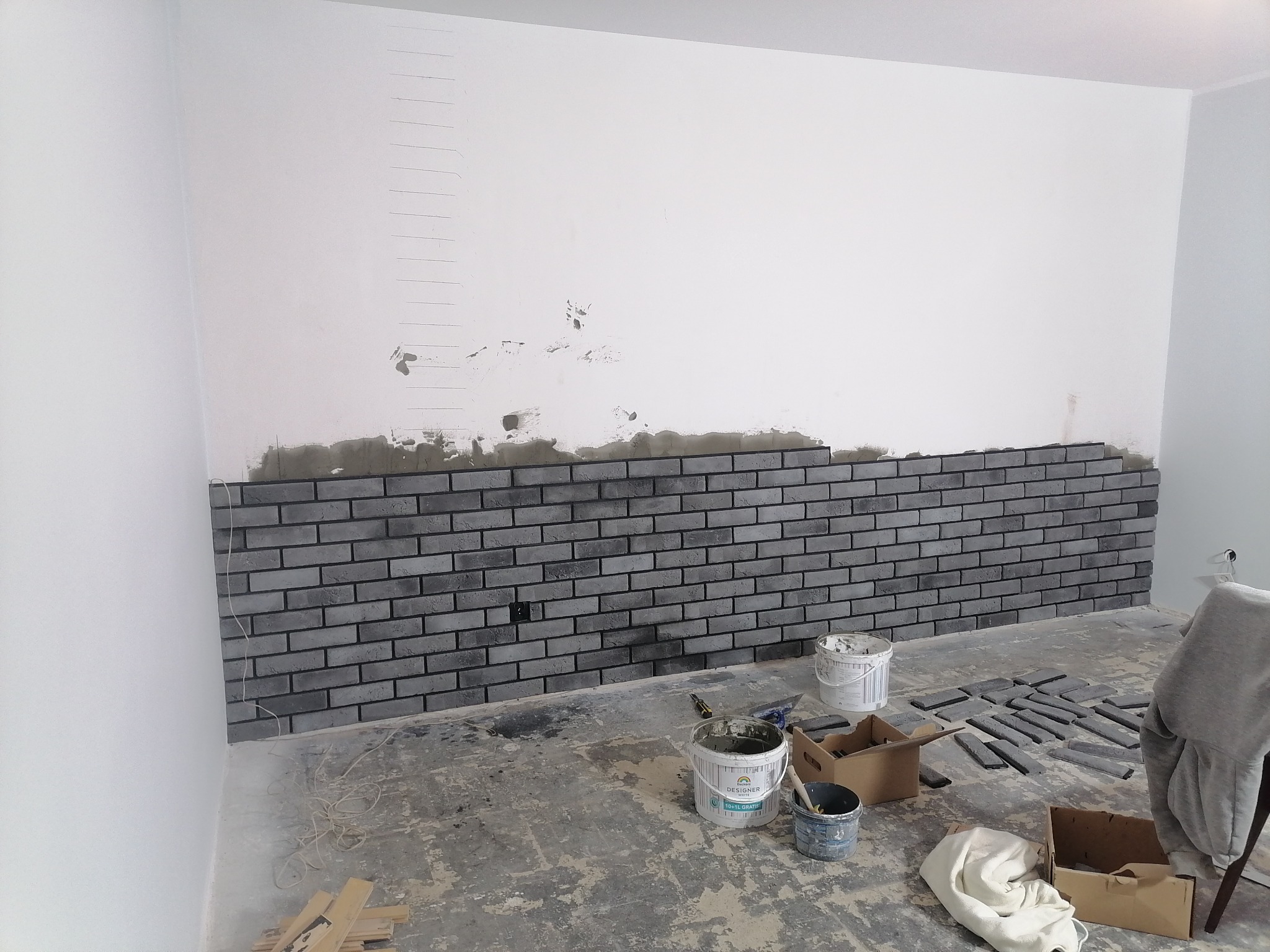 Montaż dekoracyjnej ściany z szarej cegły na klej, widoczne wiaderka z zaprawą, poziomica i kartony z cegłami na podłodze w trakcie realizacji.
