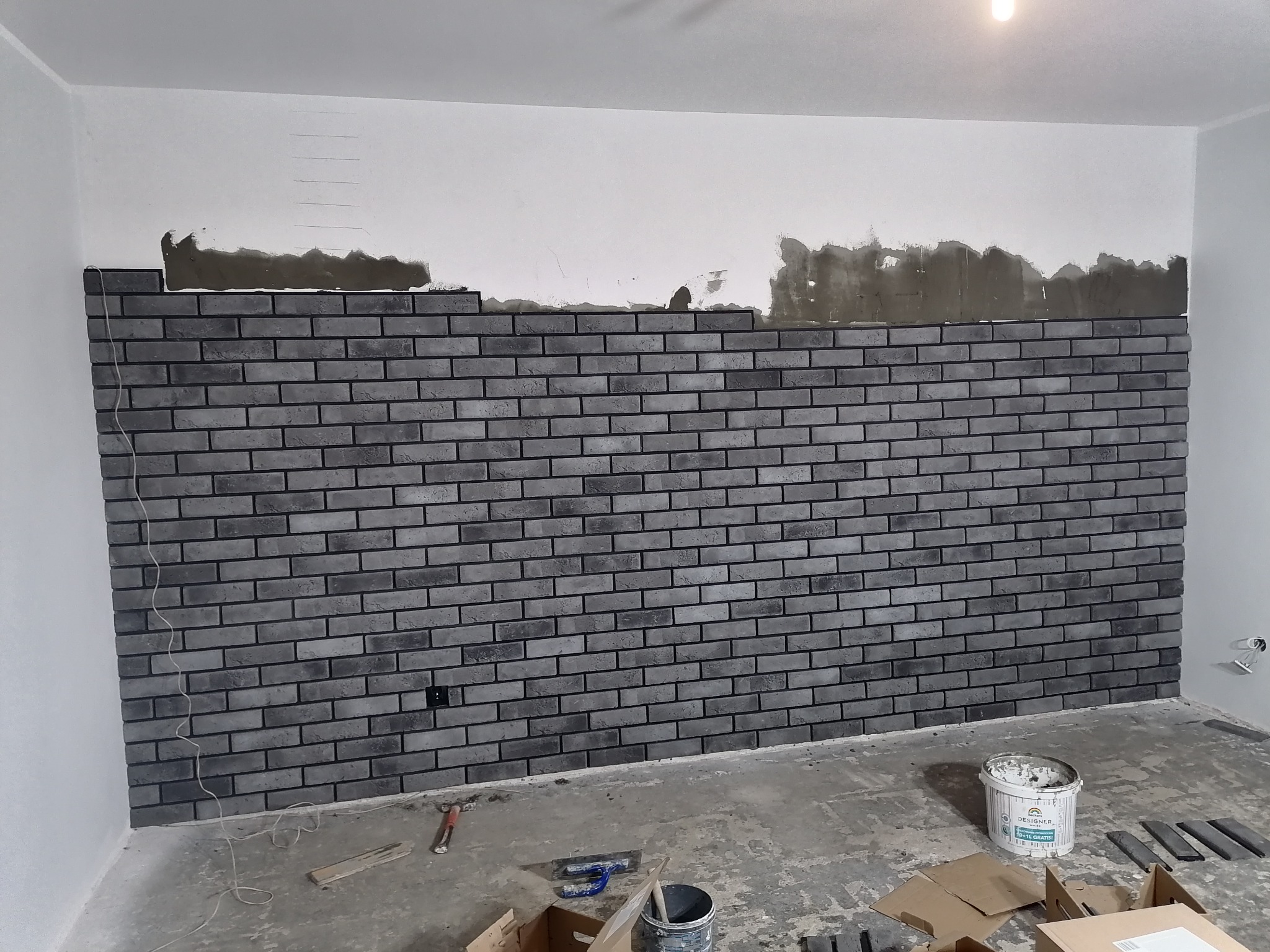 Montaż szarej cegły dekoracyjnej na ścianie, widoczny fragment ściany z naniesionym klejem, puste wiadro po farbie i narzędzia na podłodze.
