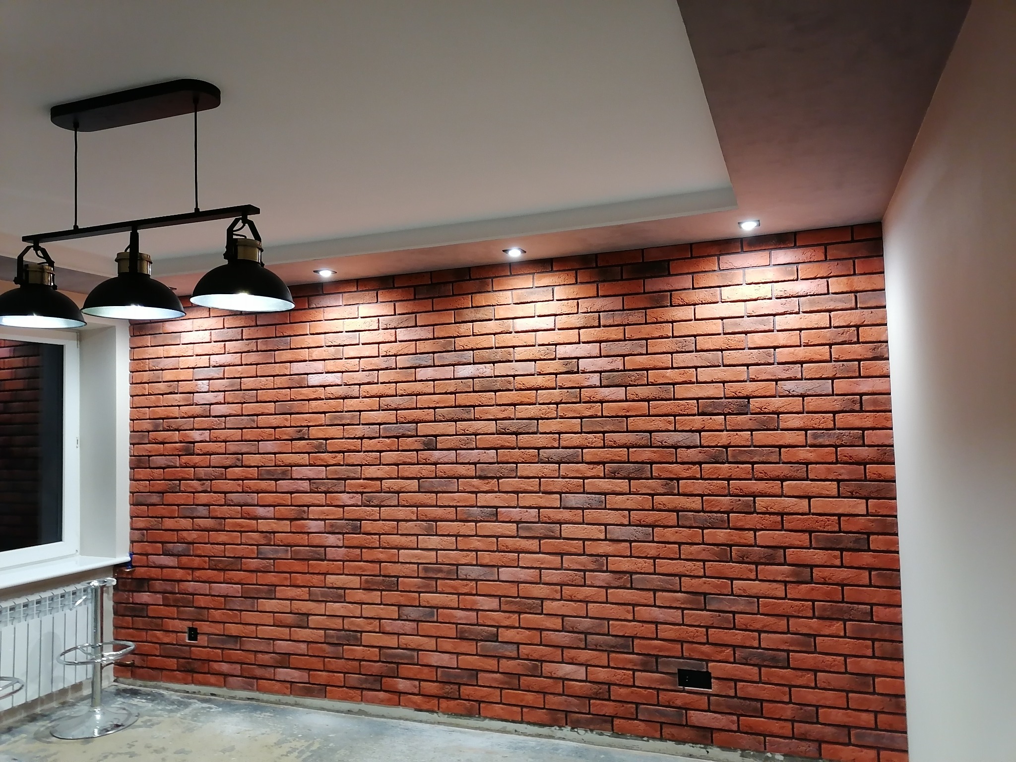 Ściana wykończona czerwoną cegłą klinkierową, oświetlona trzema lampami wiszącymi w stylu industrialnym i punktowymi reflektorami w suficie podwieszanym.