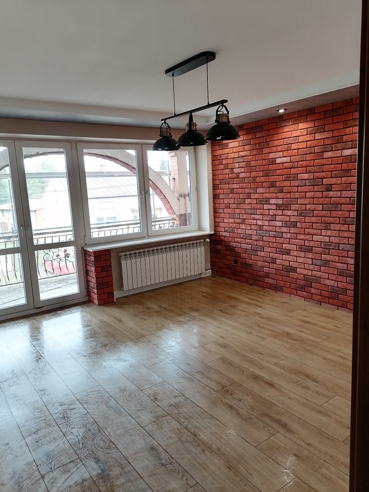 Pokój po remoncie z ceglaną ścianą, drewnianą podłogą i trzema czarnymi lampami wiszącymi, widoczne okno balkonowe i grzejnik.